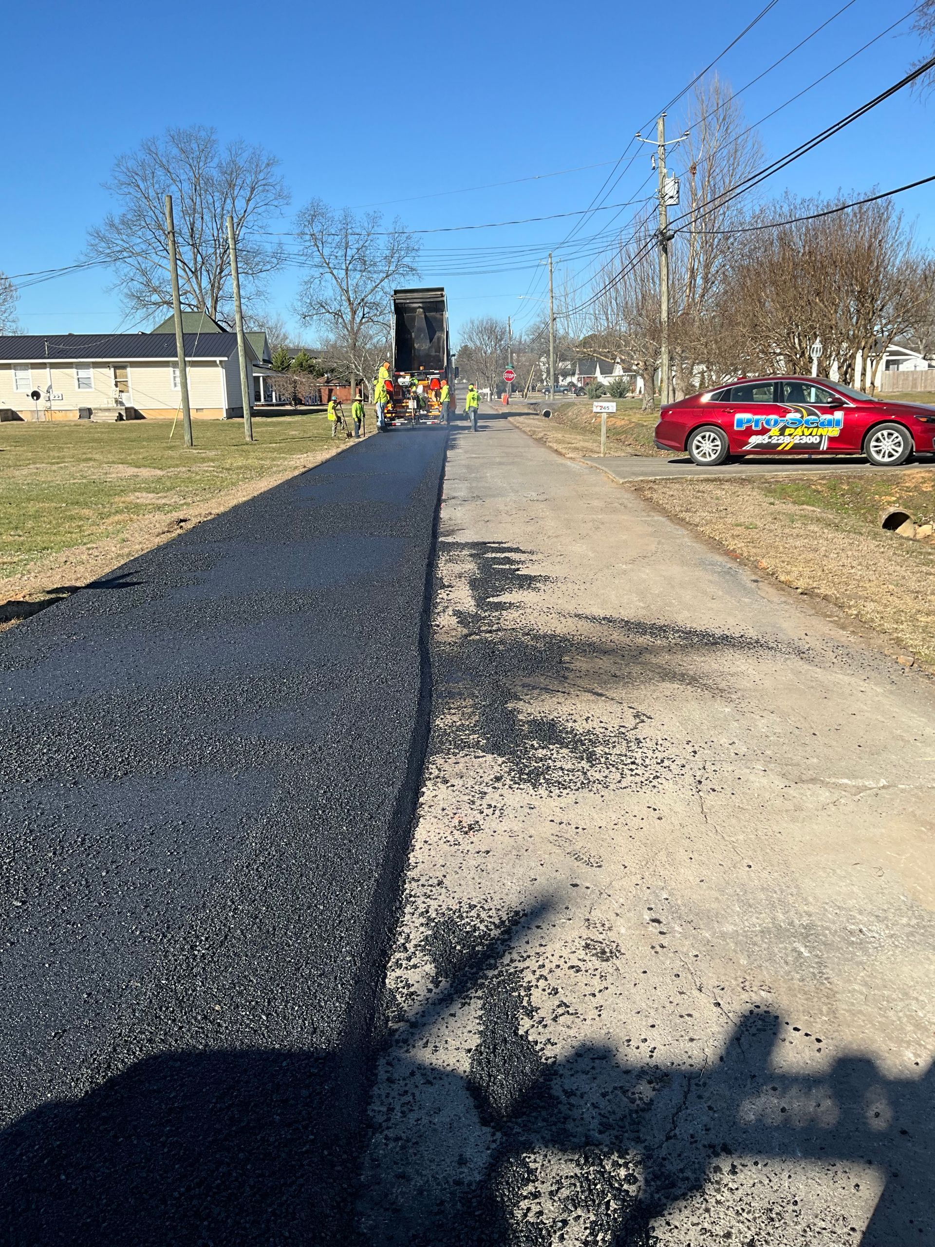 Asphalt paving a road, with a dump truck and a red car parked on the side.