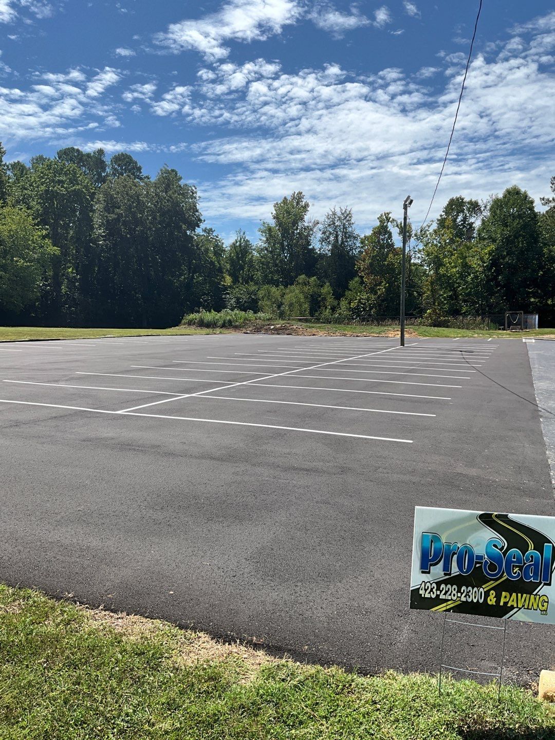 Paved parking area with white painted parking spaces, surrounded by grass, trees, and blue sky with clouds.
