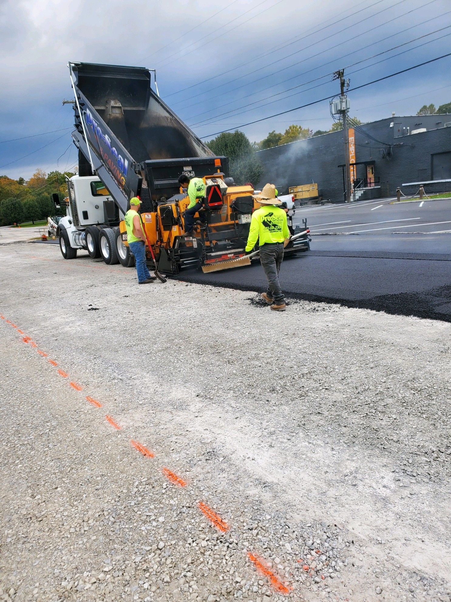 Asphalt paving in progress: dump truck unloading, paver machine laying blacktop. Workers in vests and hard hats.