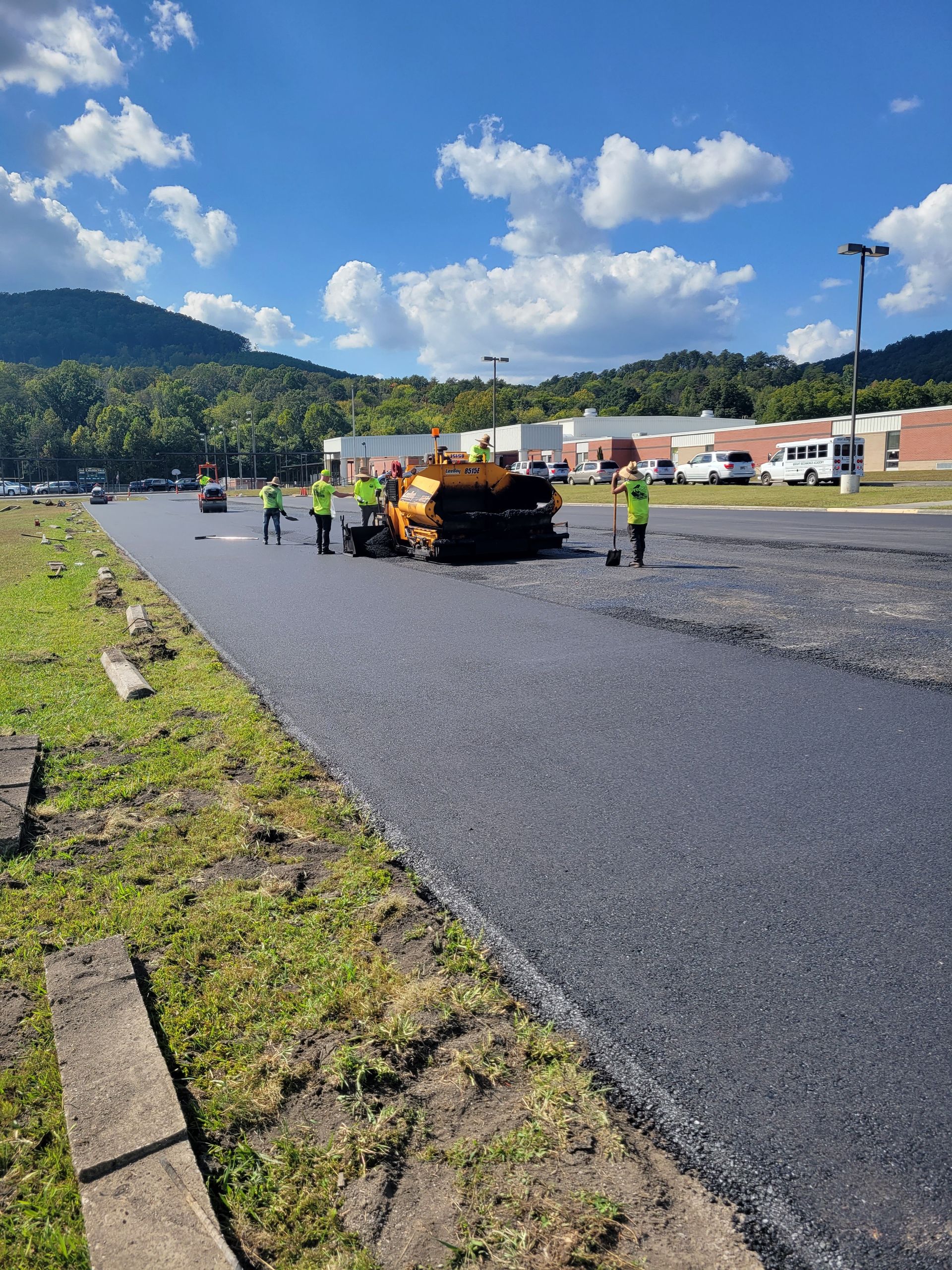Workers paving a road with a machine. The dark asphalt contrasts with the green grass. Bright blue sky with clouds.