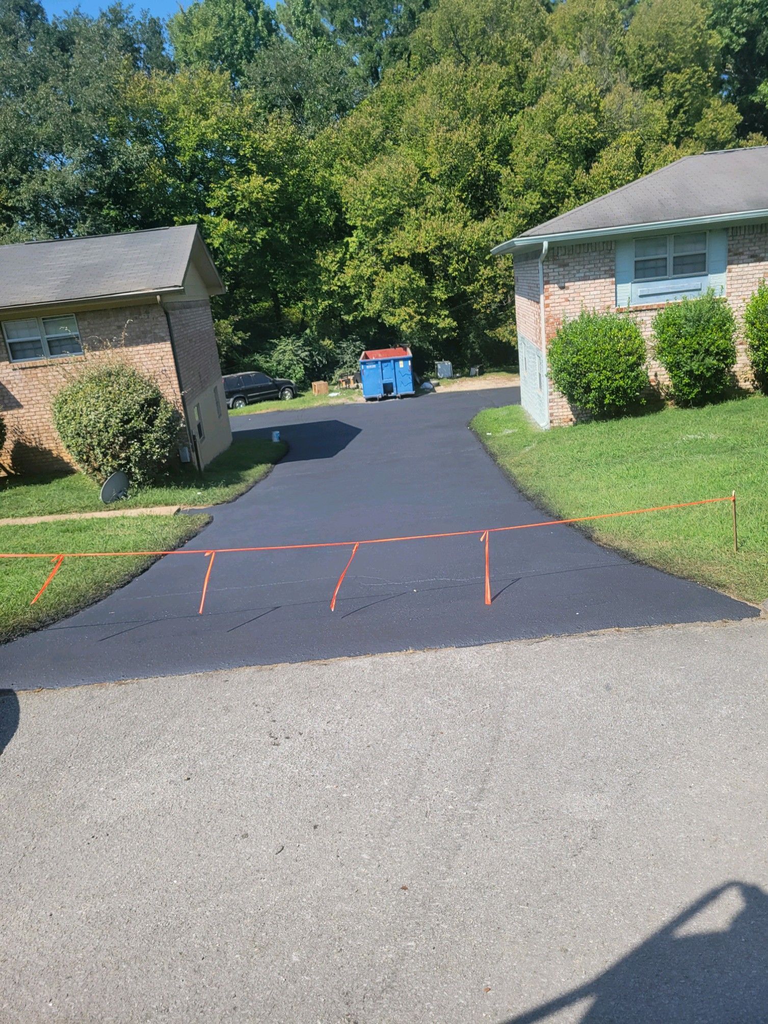 New black asphalt driveway between two brick houses, with orange marking tape.