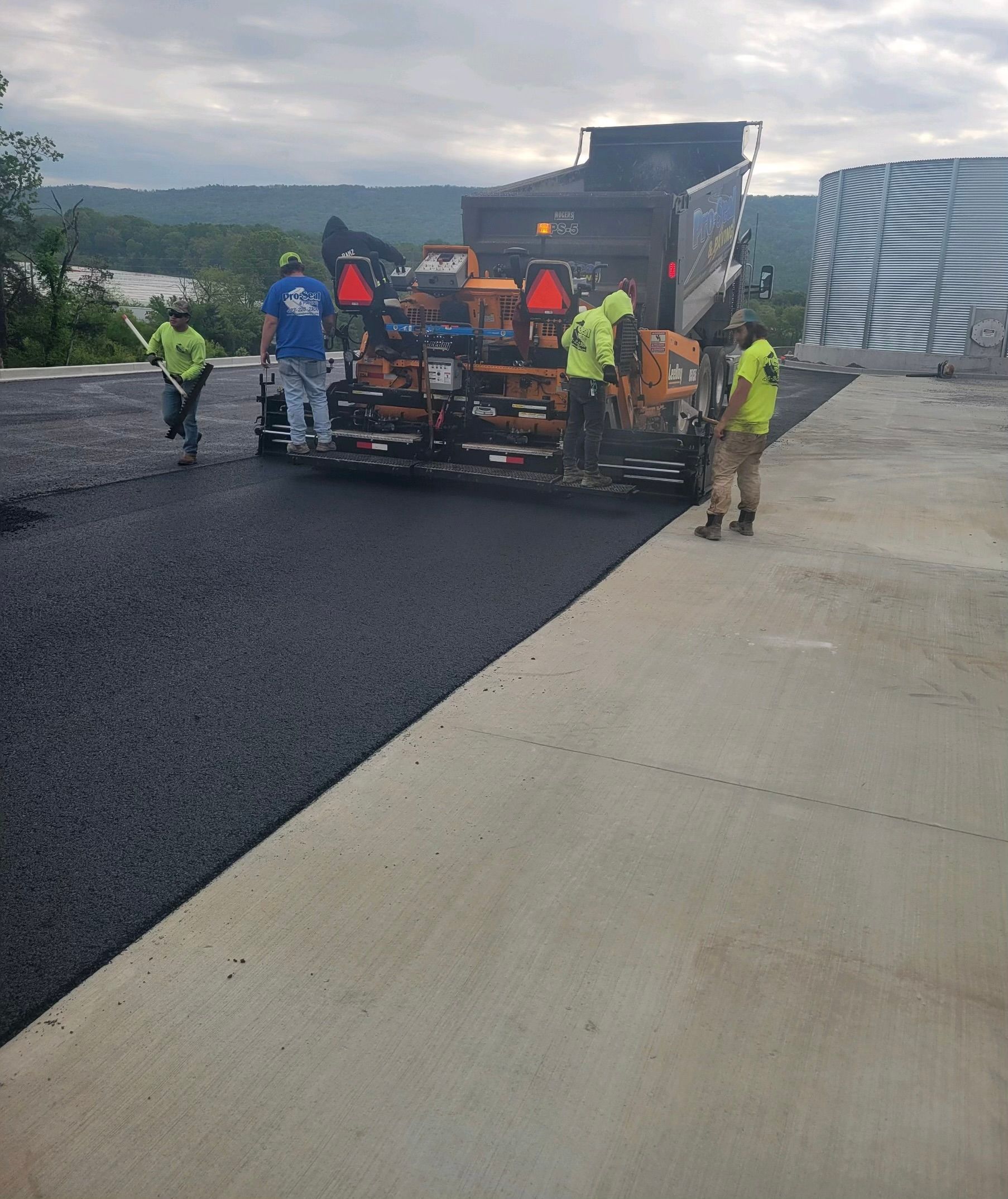 Road paving: Asphalt paving machine laying black asphalt on concrete surface, construction workers in safety vests.