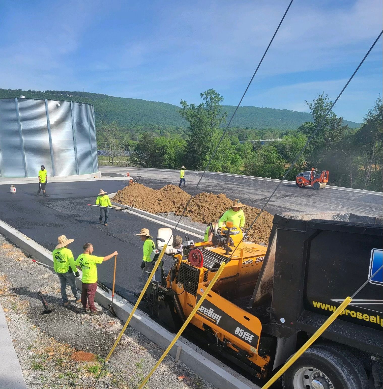 Road construction: Workers in safety vests, asphalt paving, yellow machine, dark truck, mountains in background.