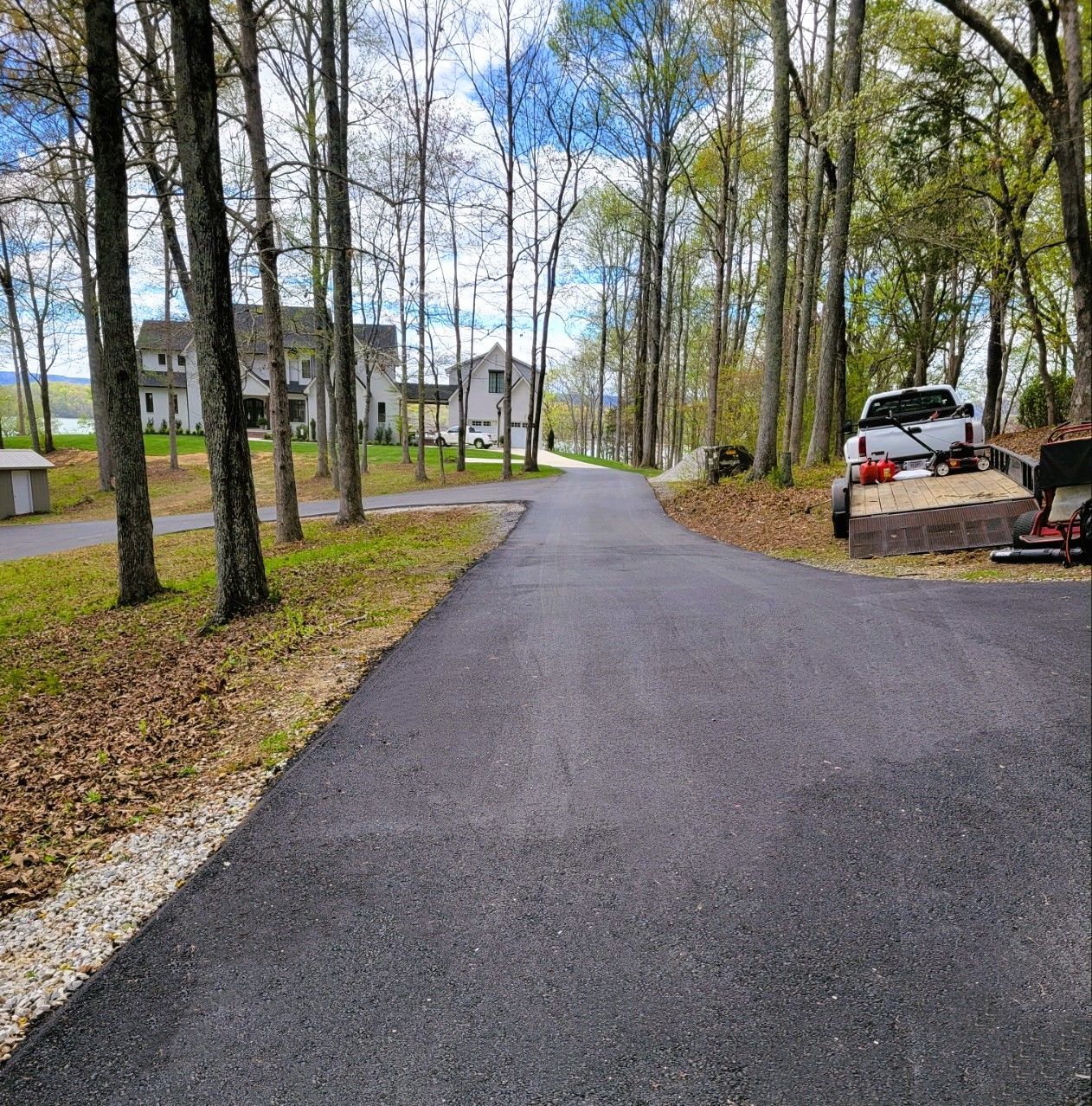 Asphalt driveway through a wooded area leading to houses. A truck and trailer are on the side.