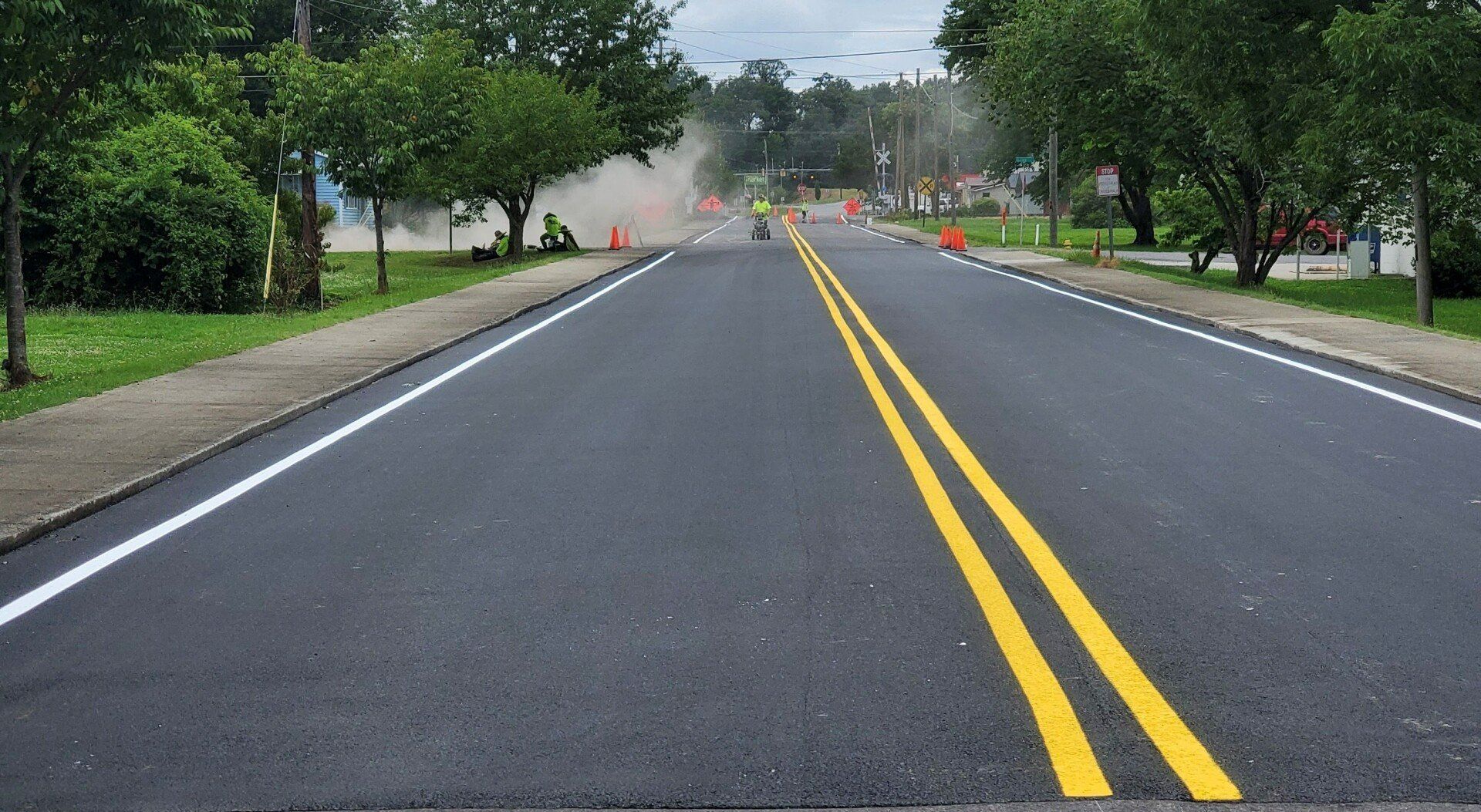 Newly paved road with double yellow lines, white edge lines, orange cones, and construction activity in the distance.