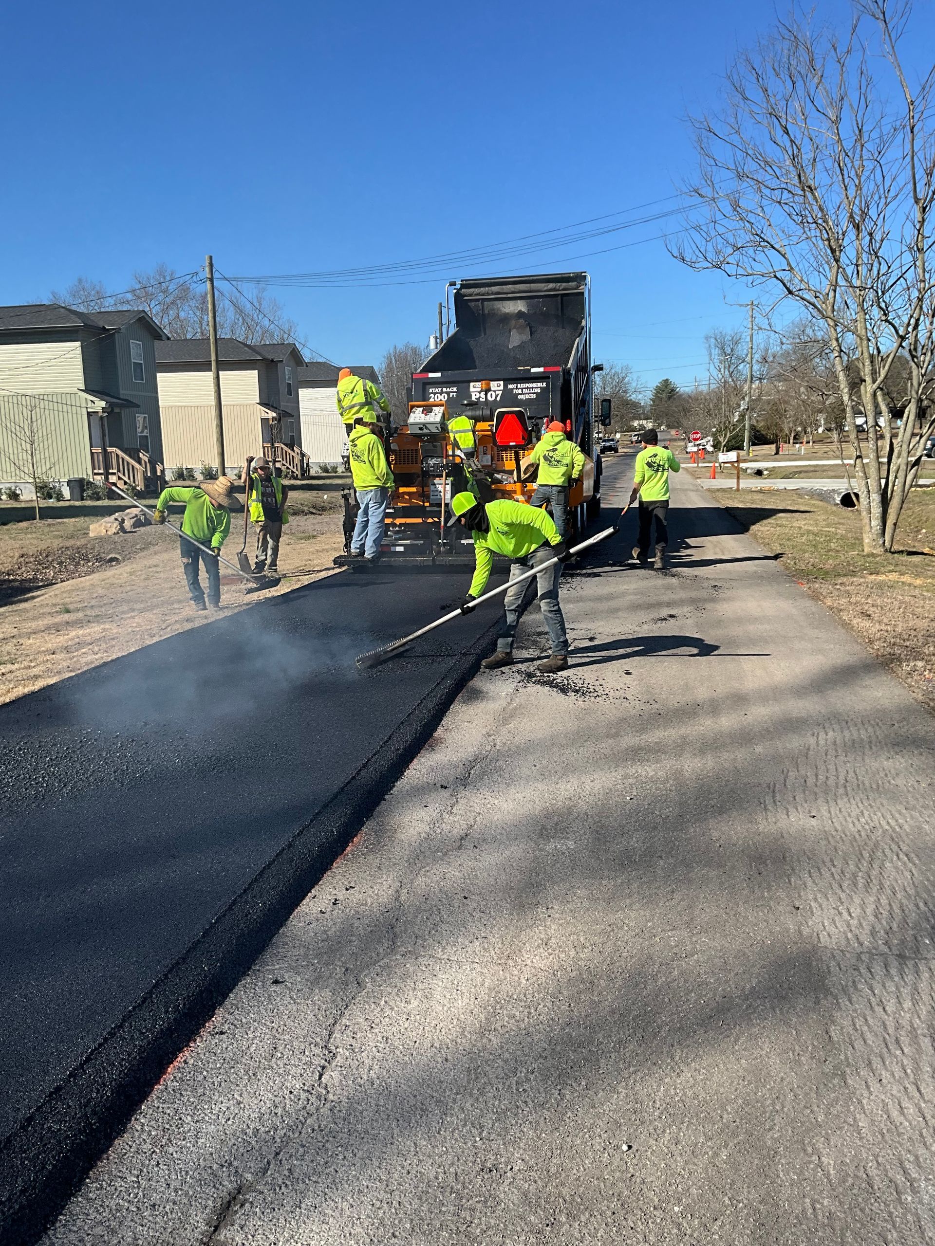 Road paving in progress. Workers in safety vests and hard hats. Paving truck laying asphalt on the street. Sunny day.