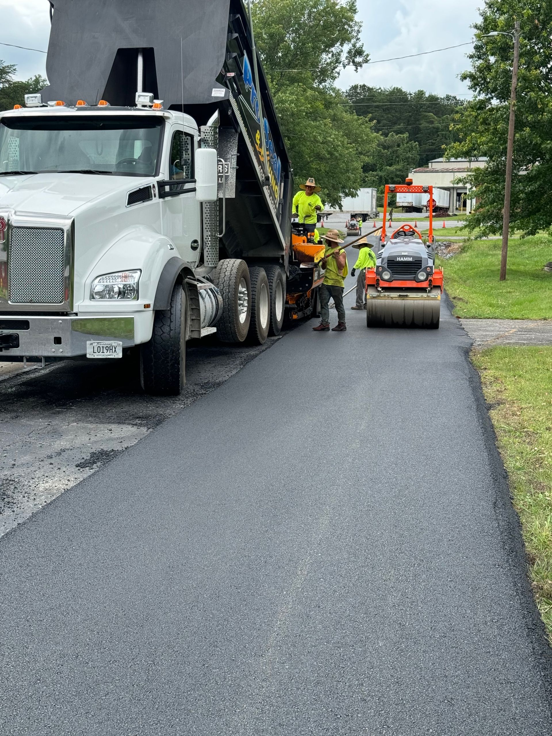 Road paving: Dump truck depositing asphalt, workers in vests, a roller compacting.