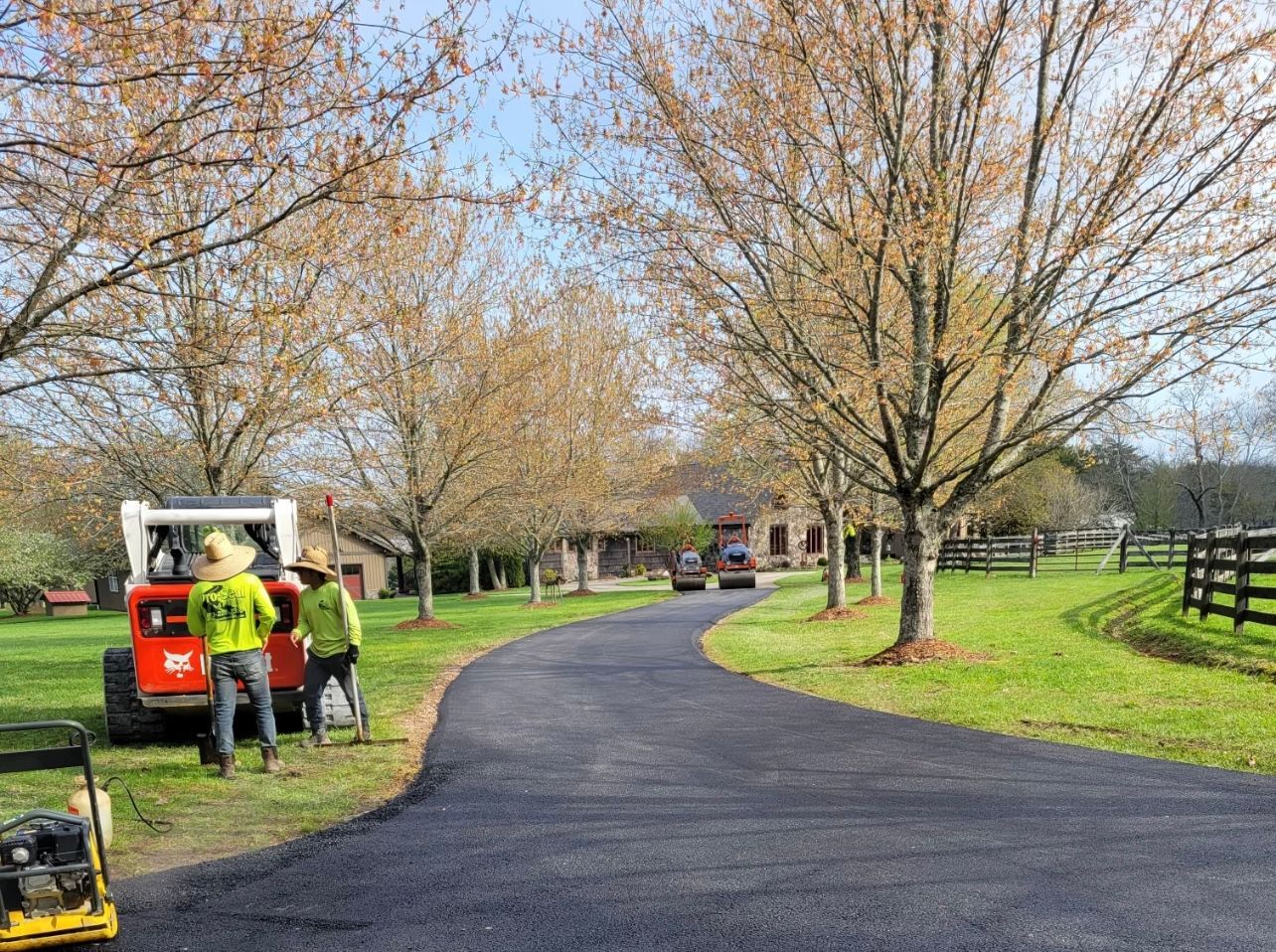 Workers paving a long, curved driveway lined with trees in a grassy setting.
