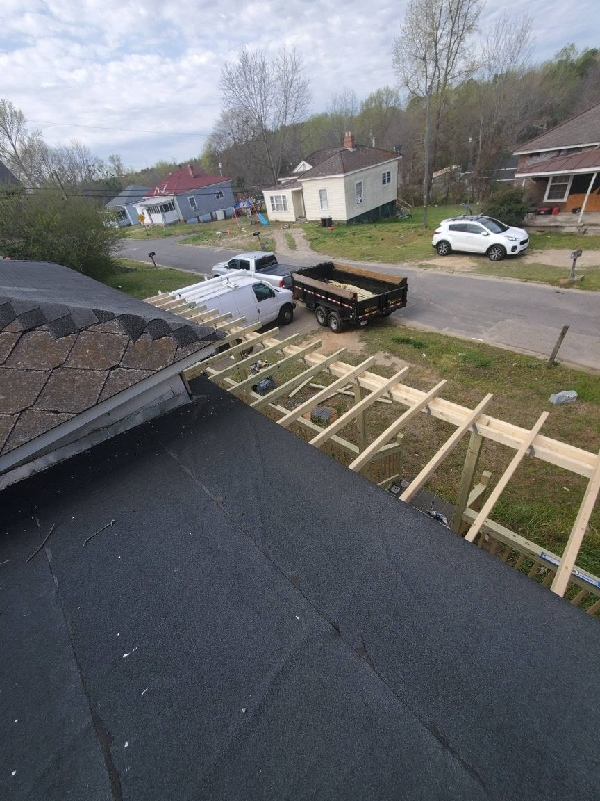 Roof construction with exposed beams. A truck and trailer are nearby. Houses and a road in the background.