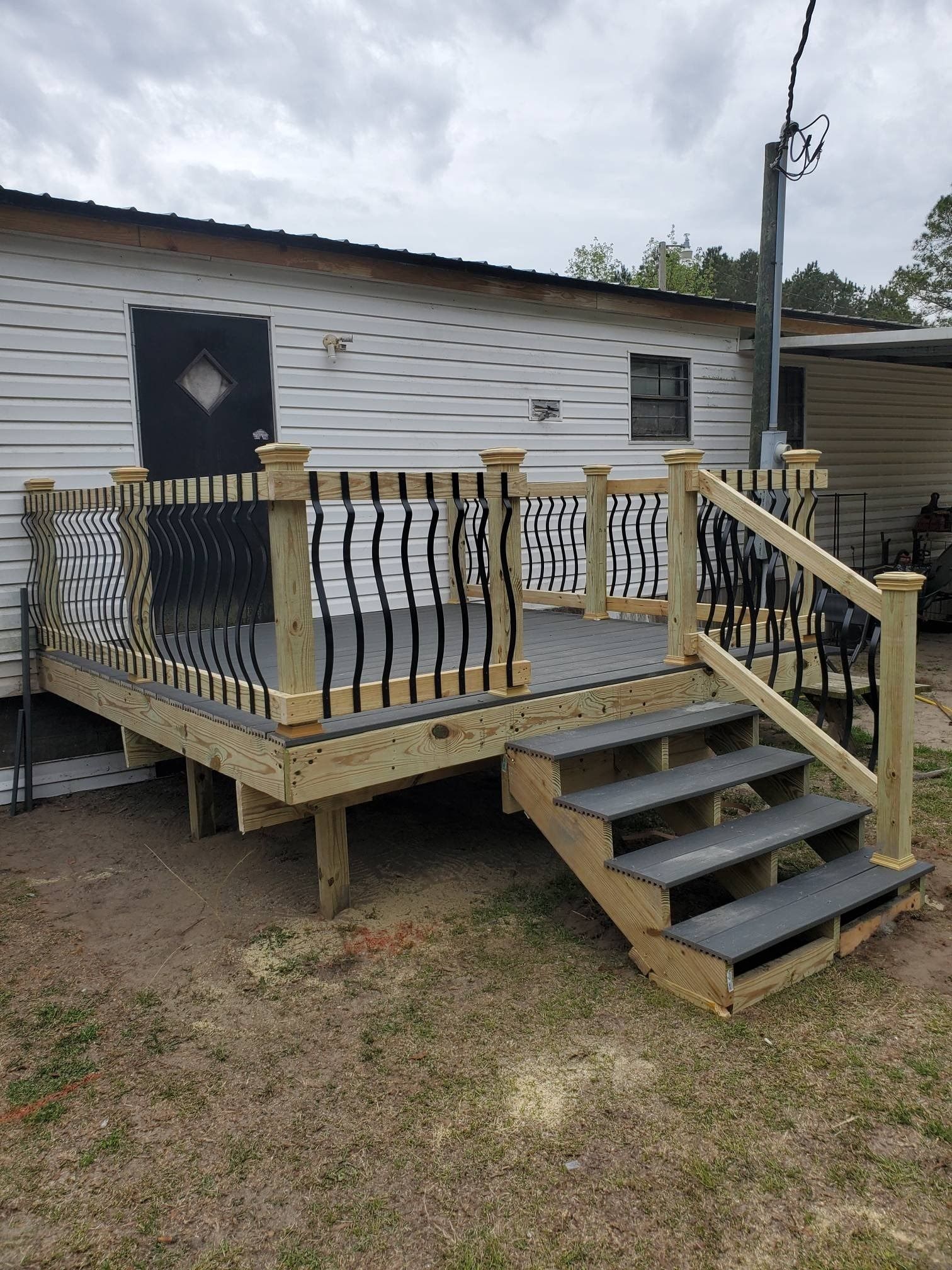 Wooden deck with steps, black railing, and gray decking, next to a white house with a black door.