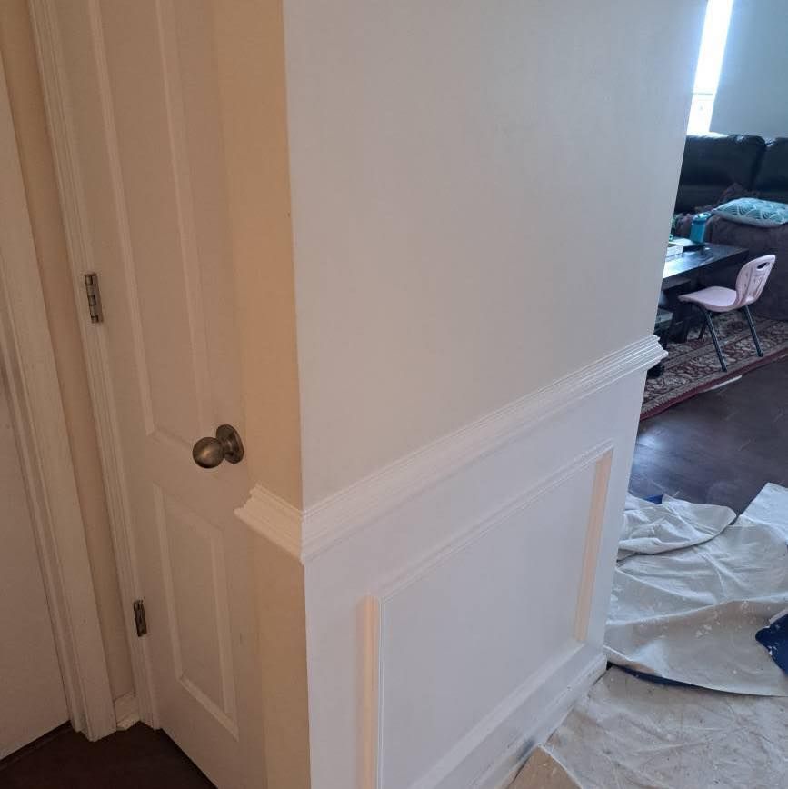 White painted wall with molding, door, and a glimpse into a living room.
