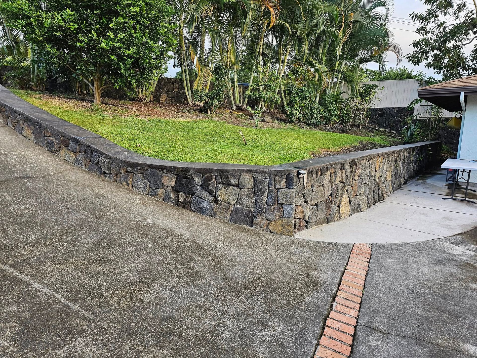Curved stone retaining wall beside a grassy slope and concrete patio with a brick strip in front