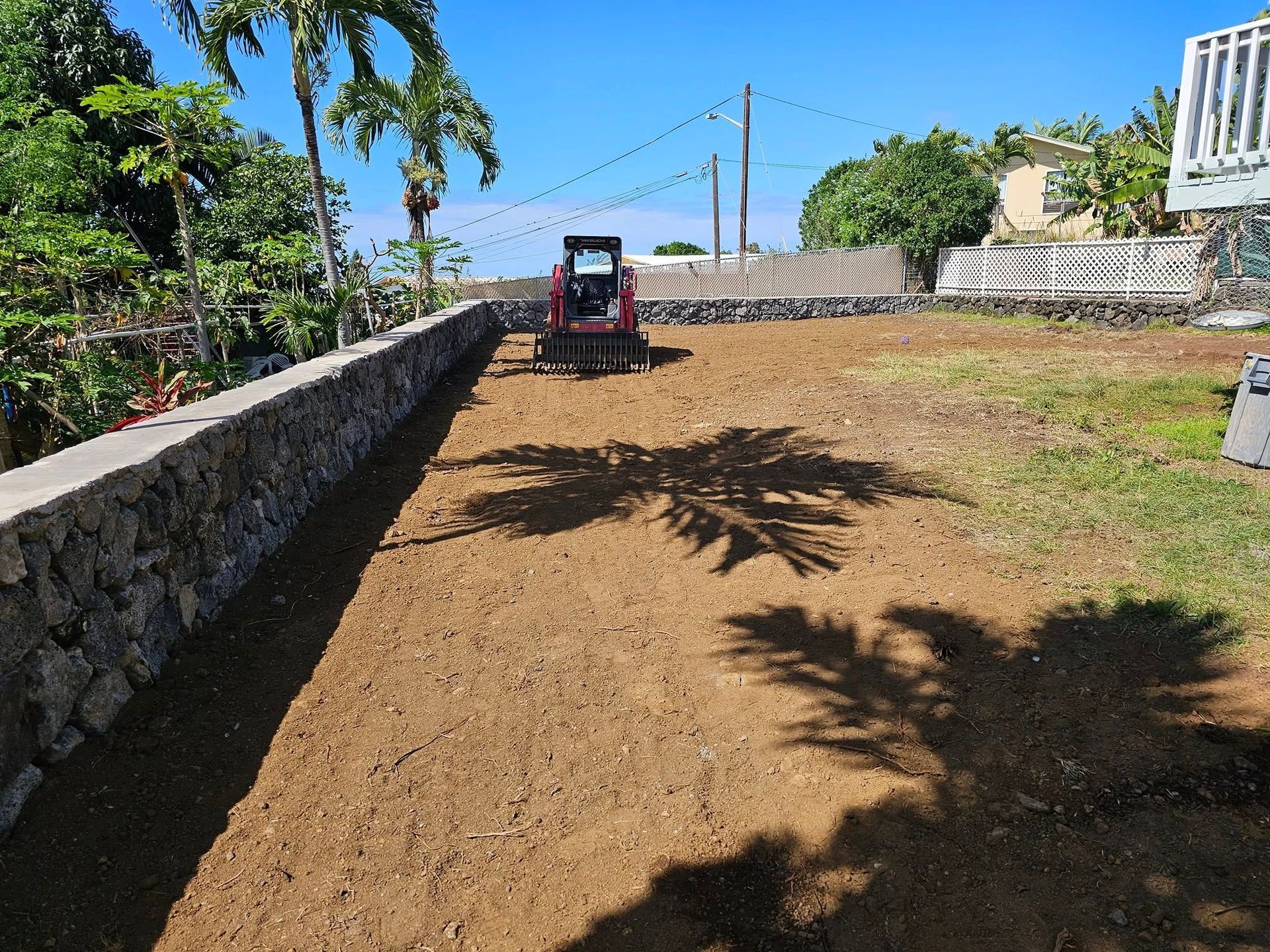 Small tractor leveling a dirt yard beside a stone wall under a clear blue sky