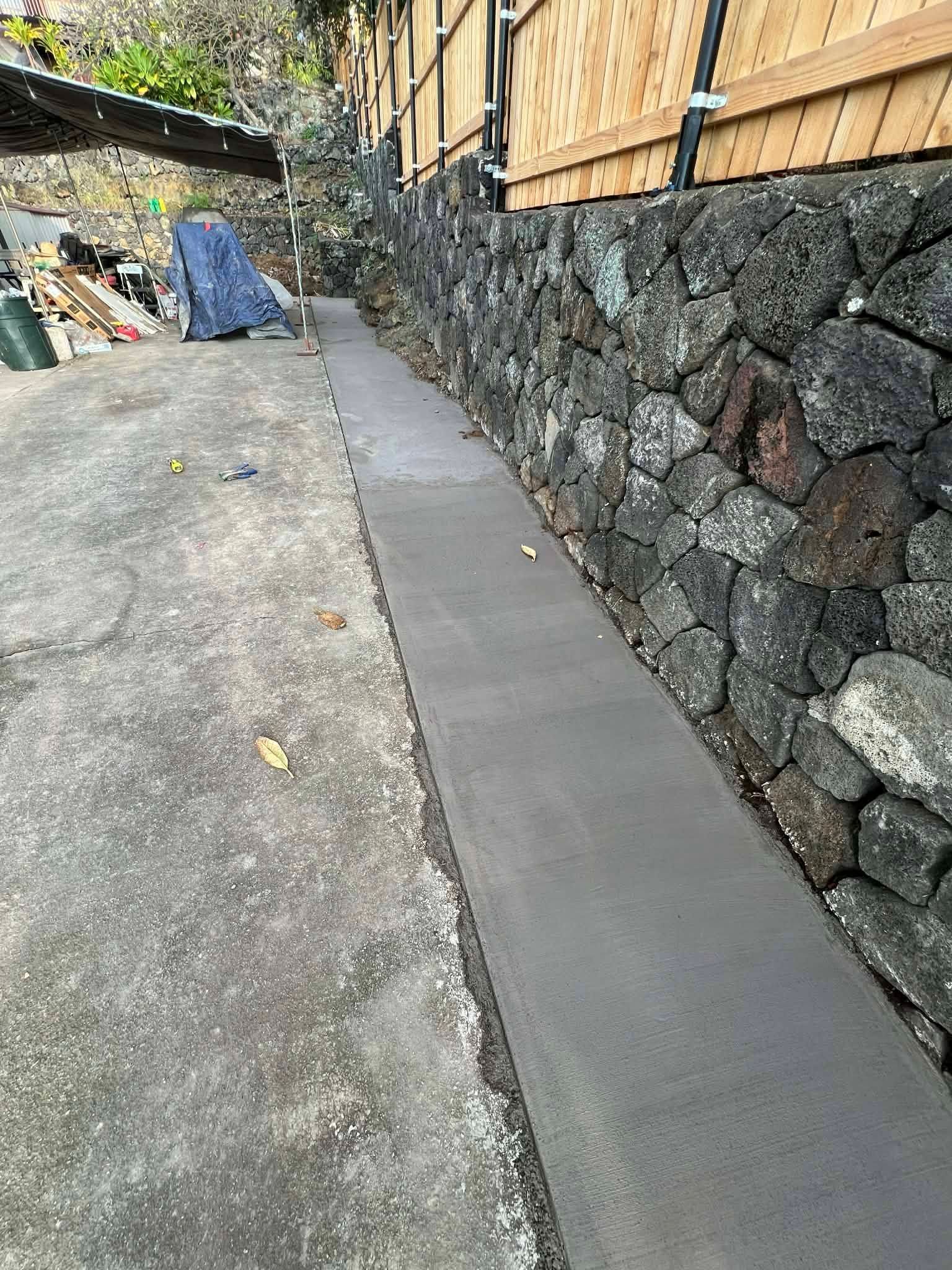 Narrow concrete walkway beside a stone retaining wall and wooden fence, with a gravel driveway on the left.