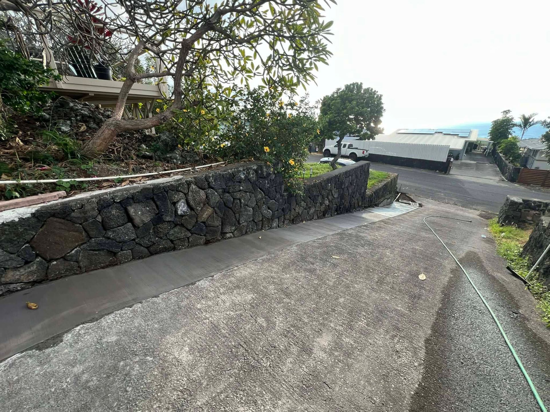 Stone retaining wall beside a sloped concrete path, with trees and ocean-view parked cars ahead