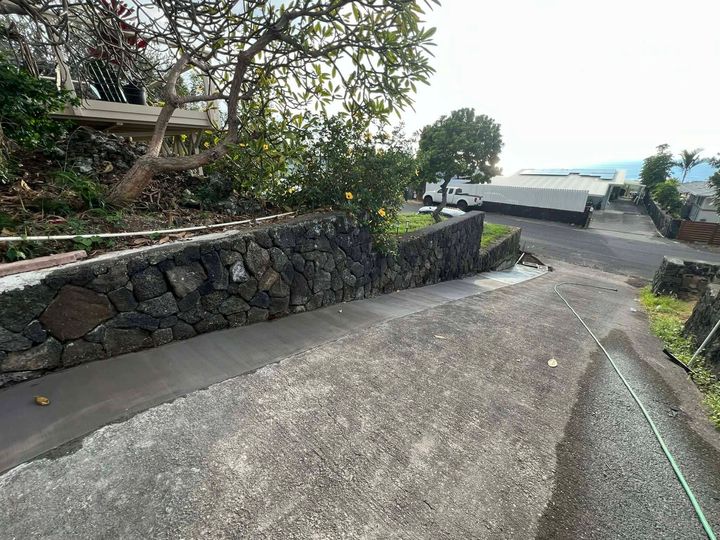 Stone retaining wall beside a sloped concrete path, with trees and ocean-view parked cars ahead