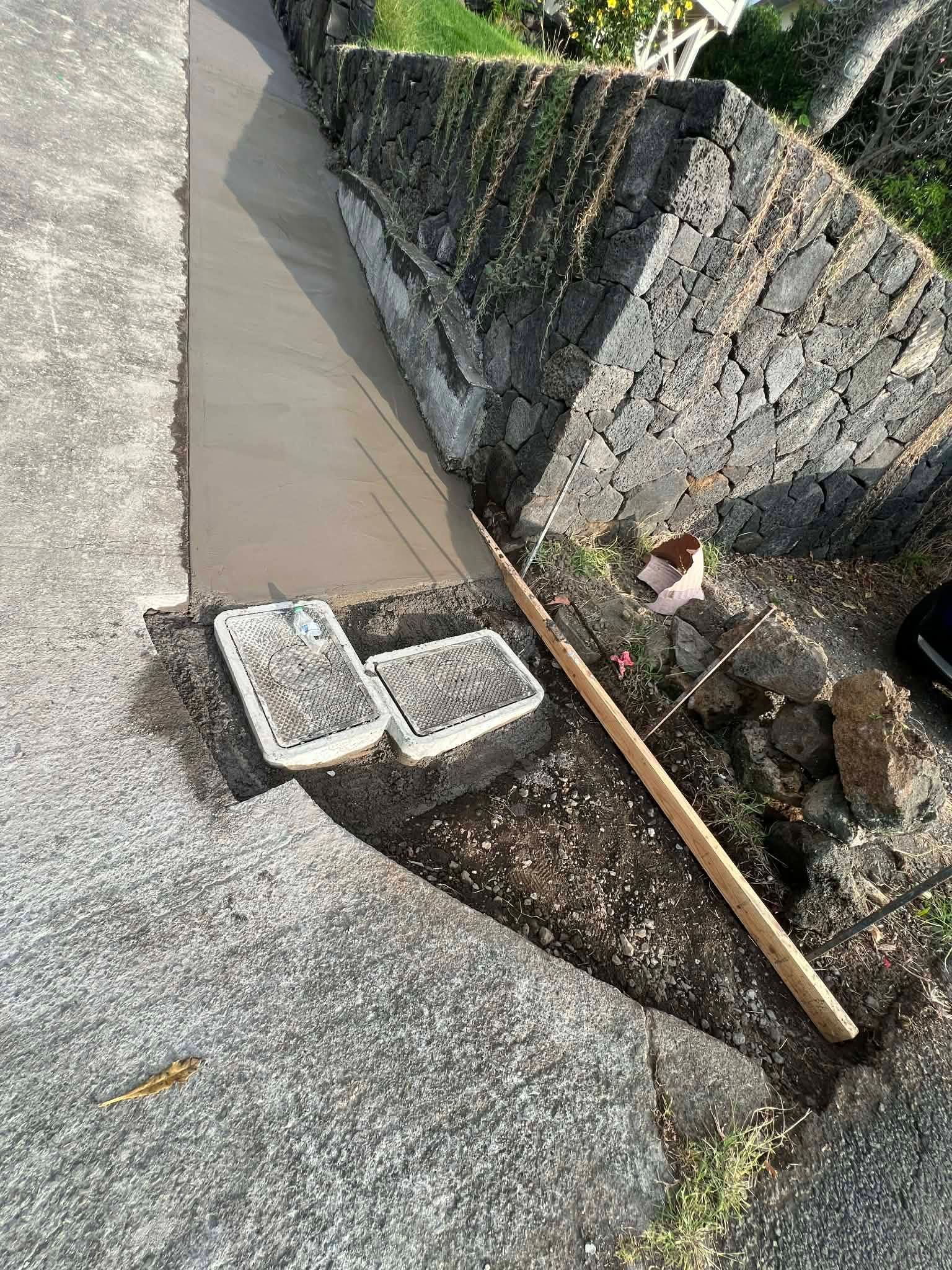 Damaged roadside ditch with cracked concrete, two loose cinder blocks, and a wooden stick on gravel.