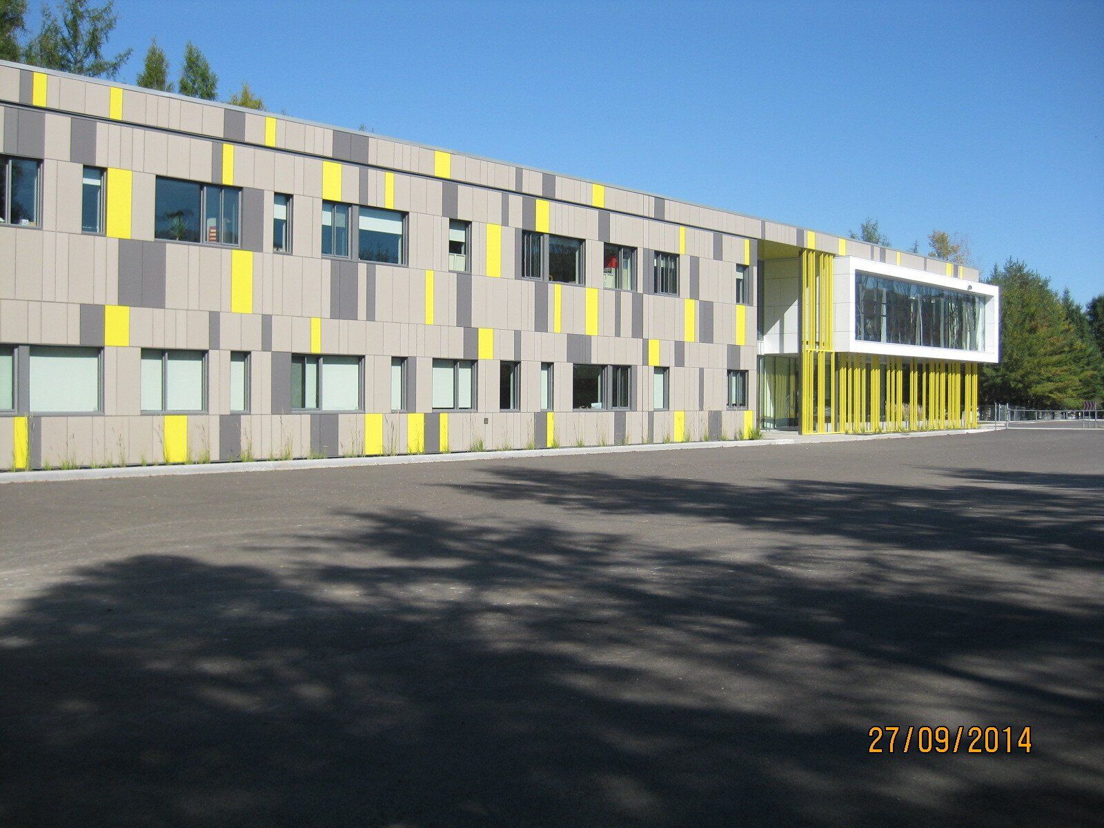 Bâtiment moderne à la façade grise et jaune, fenêtres rectangulaires et entrée vitrée. Journée ensoleillée.