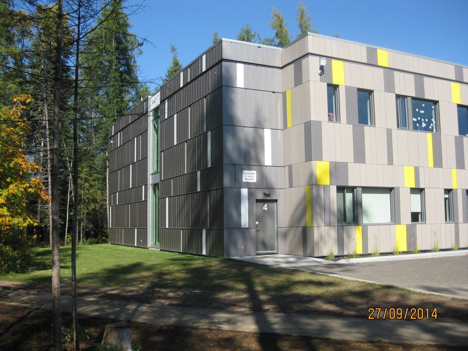 Bâtiment moderne aux panneaux et fenêtres gris, jaunes et blancs, entouré d'arbres par une journée ensoleillée.