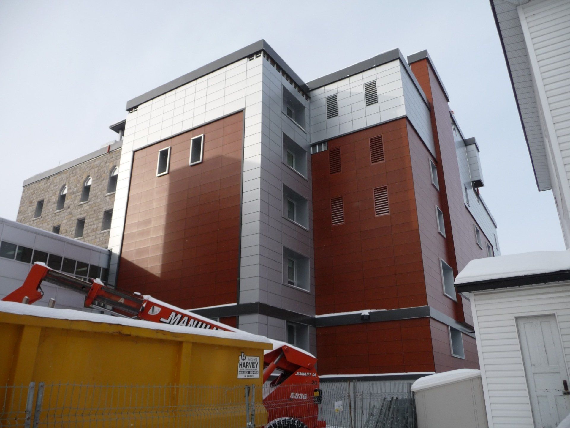 Bâtiment moderne aux panneaux rouges et argentés, avec de la neige et des engins de chantier à l'avant.