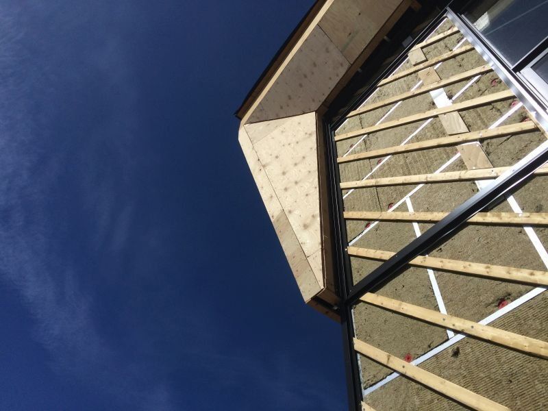 Extérieur d'un bâtiment en construction, montrant la charpente en bois, l'isolation et un ciel bleu dégagé.