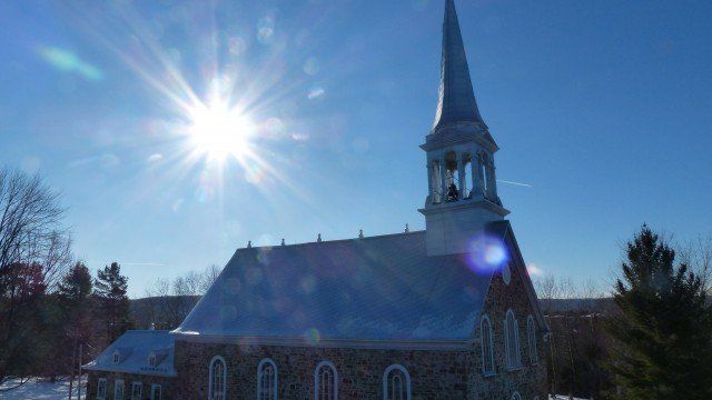 Église en pierre au haut clocher, baignée d'un soleil éclatant sur un ciel bleu, la neige recouvrant le sol.