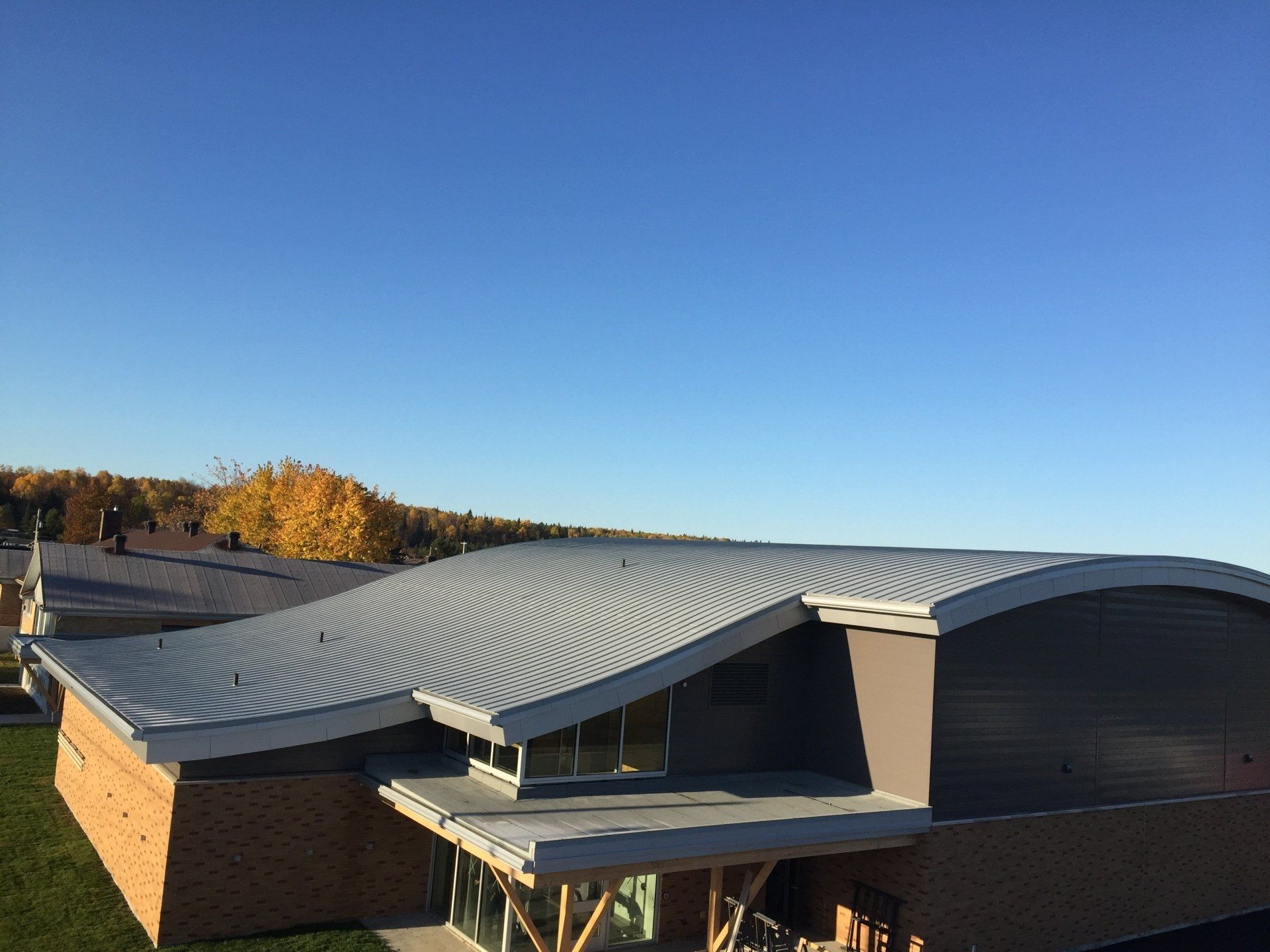 Bâtiment au toit métallique incurvé se détachant sur un ciel bleu clair, des murs de briques et des feuillages d'automne en arrière-plan.