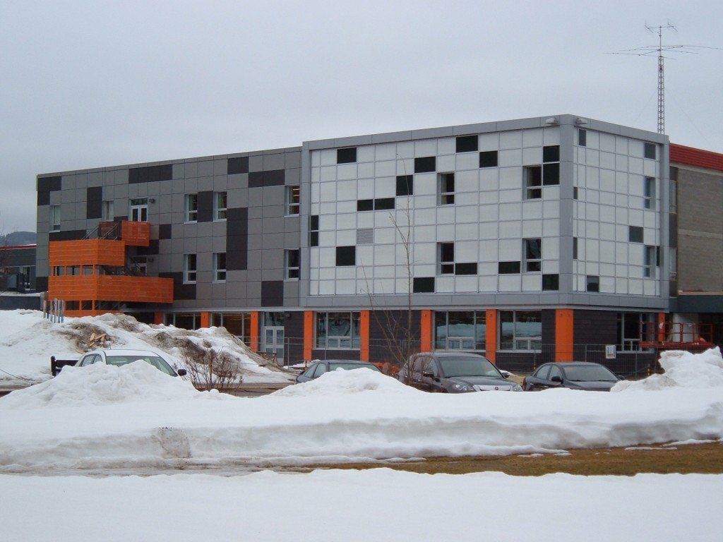 Bâtiment moderne à la façade orange et grise, sol enneigé, voitures garées devant.