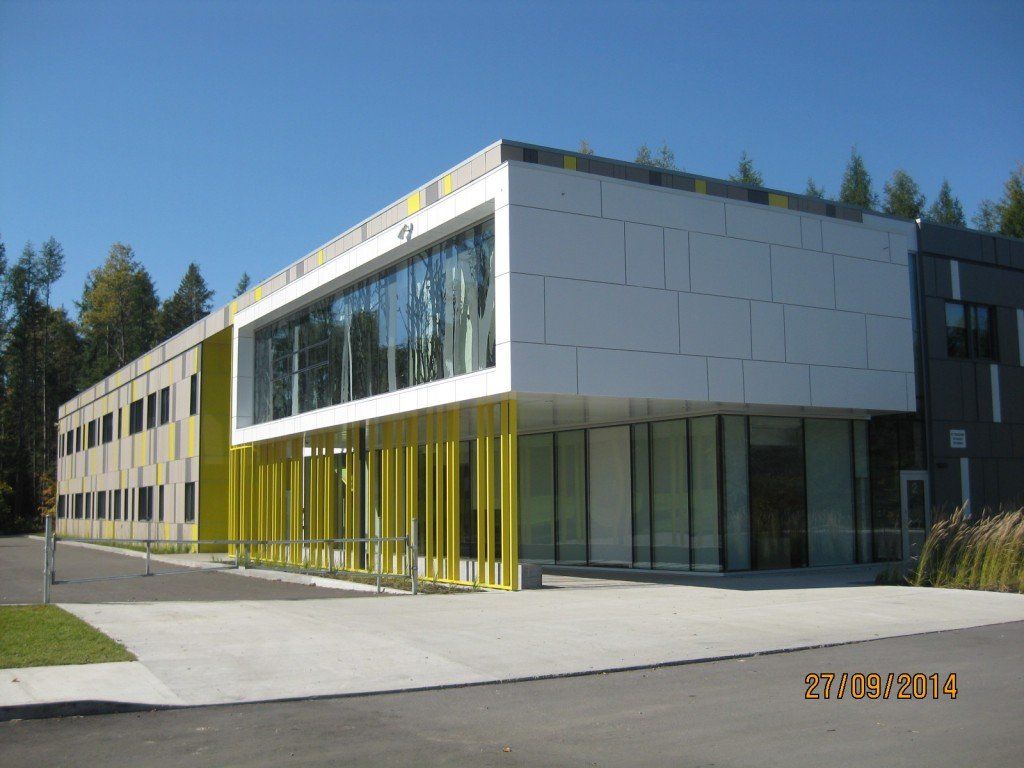Bâtiment moderne à la façade blanche et jaune, grandes fenêtres et poutres de soutien jaunes. Journée ensoleillée.