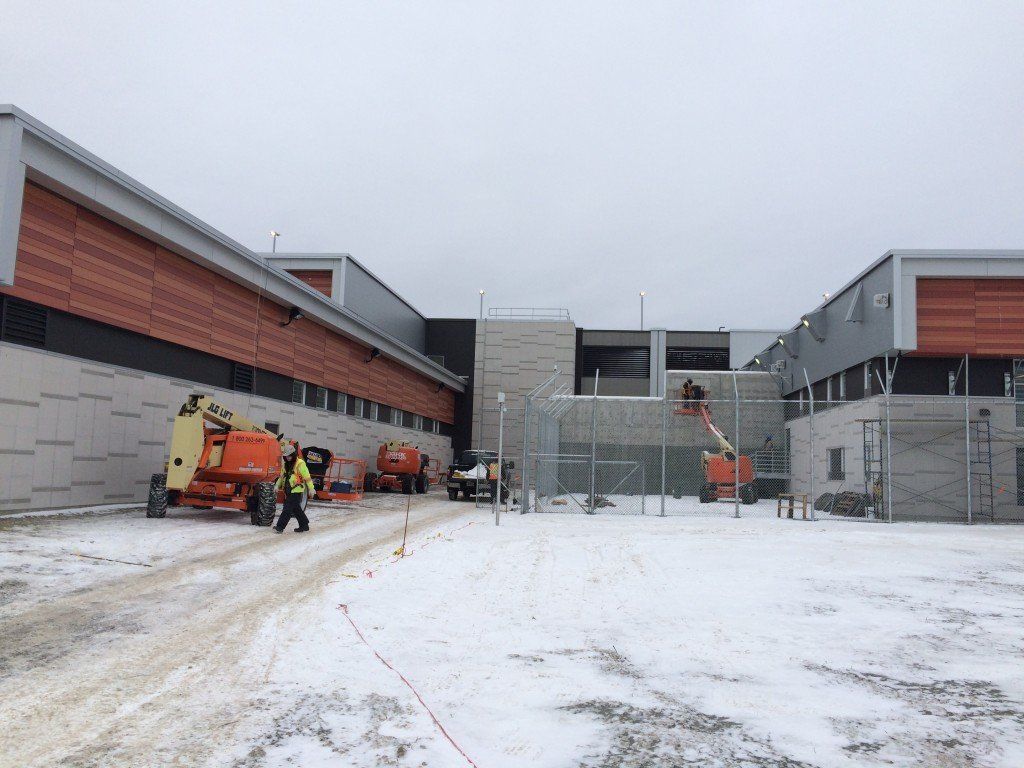 Chantier de construction enneigé avec nacelles élévatrices et ouvriers. Le bâtiment est constitué de panneaux bruns et gris.