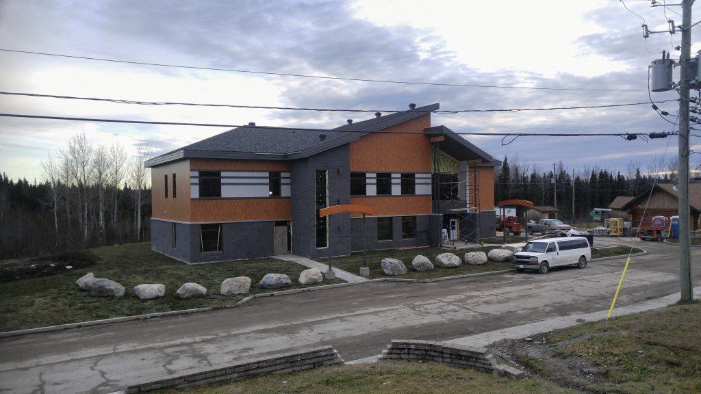 Bâtiment moderne à la façade orange et grise, ciel nuageux. Chemin de gravier, camionnette blanche, poteau électrique.