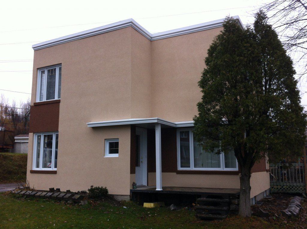 Maison beige à deux étages avec boiseries et fenêtres blanches. Grand sapin devant. Ciel couvert.
