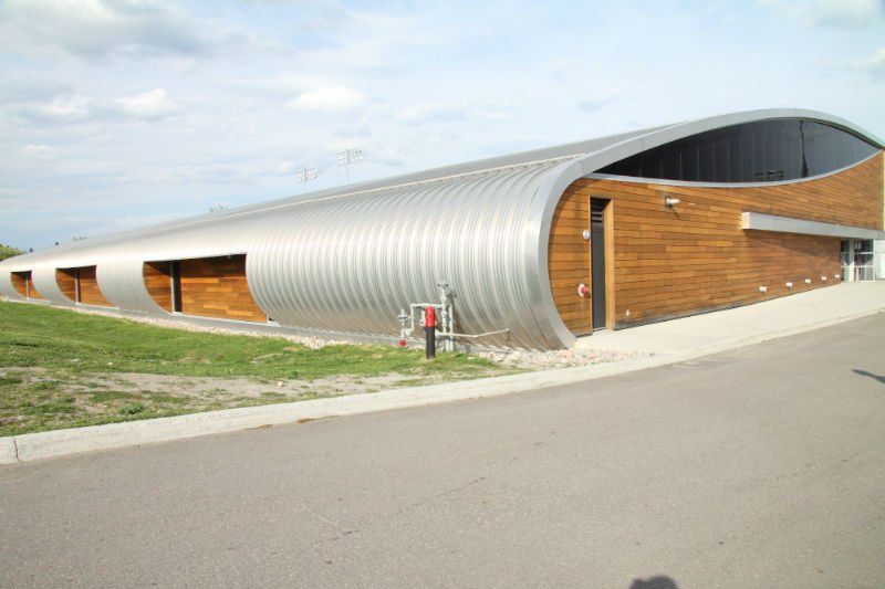 Bâtiment incurvé avec toit en tôle ondulée et éléments en bois, sur une esplanade pavée et gazonnée.