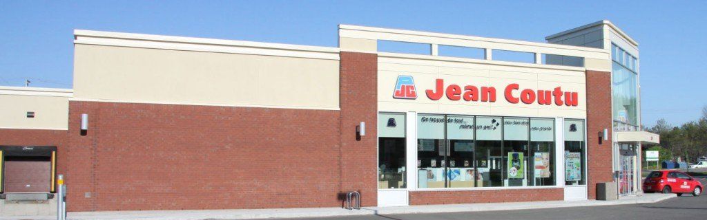 Pharmacie Jean Coutu, extérieur. Bâtiment en briques beiges avec de grandes vitrines. Voiture rouge garée devant.