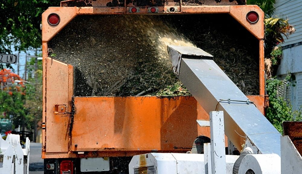 A Wood Chipper Blowing Wood Chips Into an Orange Truck Bed — Airlie Tree Specialists Pty Ltd in Proserpine, QLD