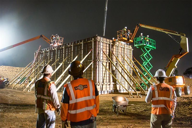 Construction workers inspecting a nighttime concrete form with cranes and a green scissor lift