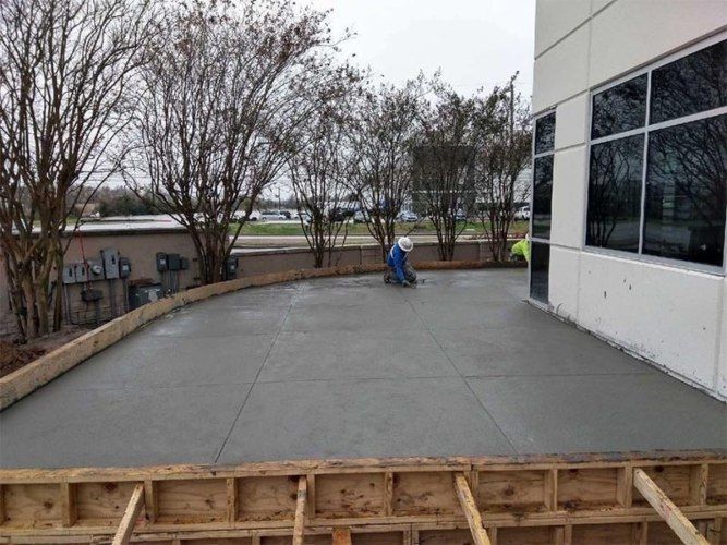 Empty concrete patio beside a building, with trees and a worker in blue crouching near the far edge.