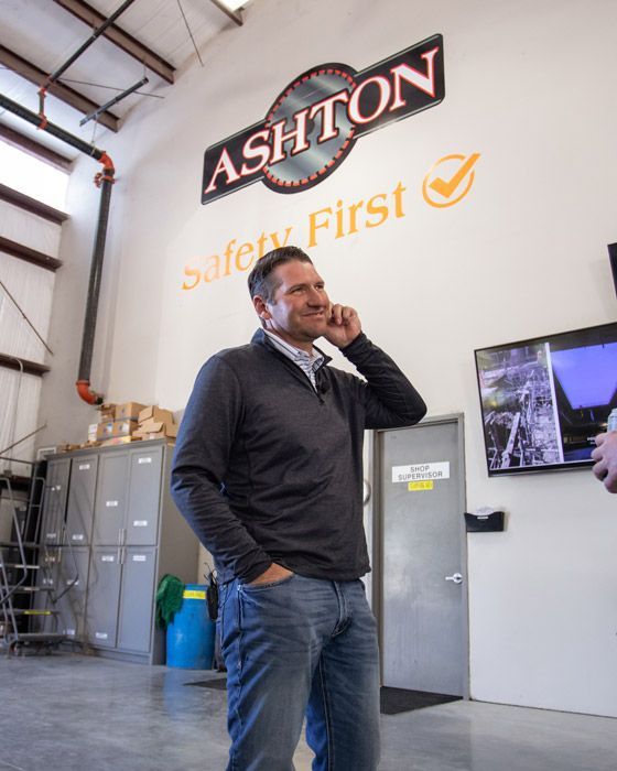 Man in dark sweater and jeans talking on a phone in an industrial office with an ASHTON Safety First sign.