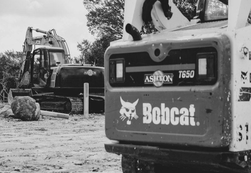 Bobcat skid-steer loader in the foreground at a construction site, with an excavator behind it.