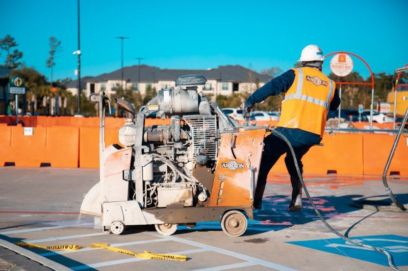 Worker in orange safety vest using a line-marking machine on a sports court outdoors.