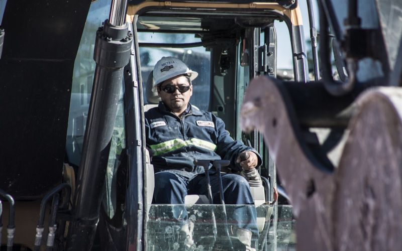 Construction worker in a cab, wearing a hard hat and sunglasses, seen through machinery.