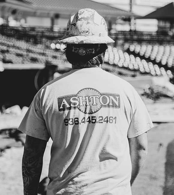Worker in a hard hat and “ASHTON” shirt walking on a stadium field, viewed from behind