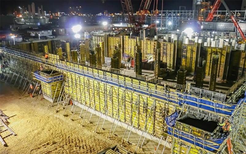 Nighttime construction site with large concrete formwork, cranes, bright floodlights, and workers below