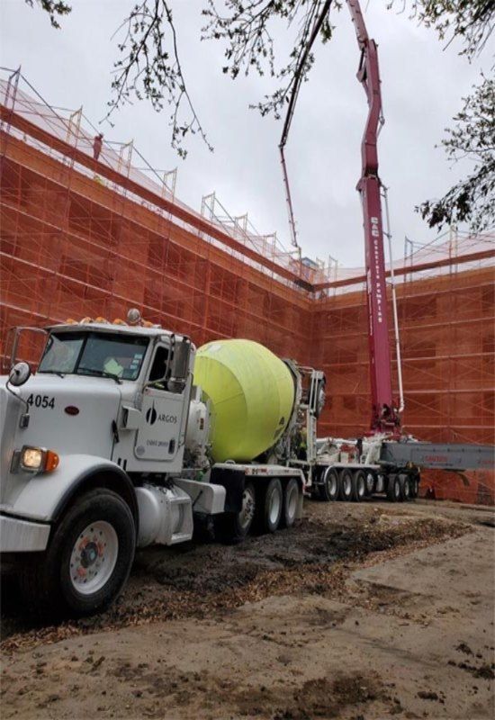 Concrete mixer truck and red boom pump at a construction site with orange safety netting.