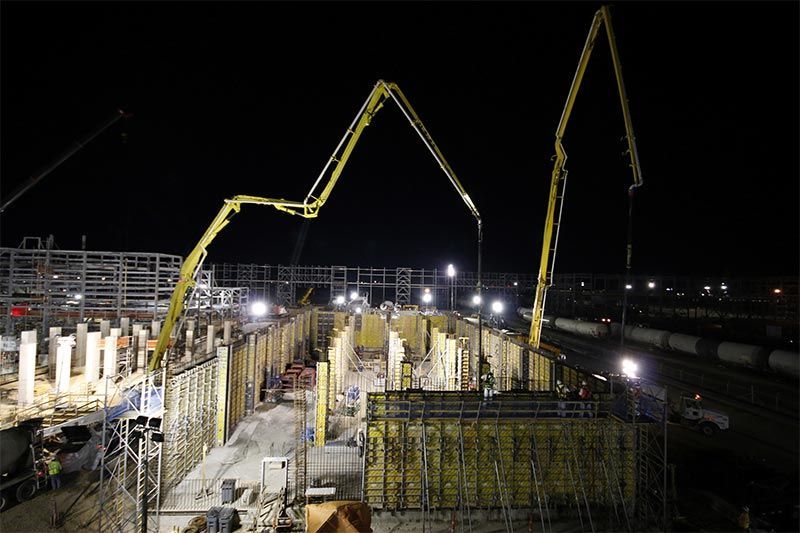 Night construction site with two concrete pump booms above rebar and formwork, lit by floodlights