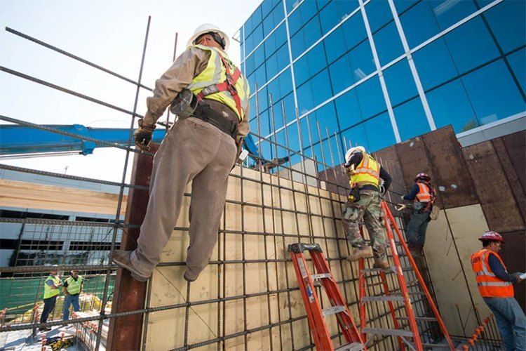 Construction workers on scaffolding beside a glass building, installing a wall panel and rebar grid.