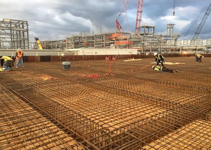 Construction workers on a rebar grid at a large building site with cranes and scaffolding.
