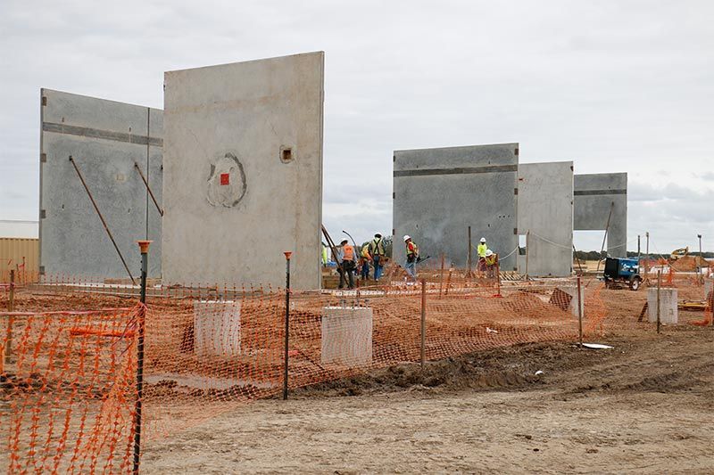 Construction site with concrete wall panels, workers, and orange safety fencing on muddy ground