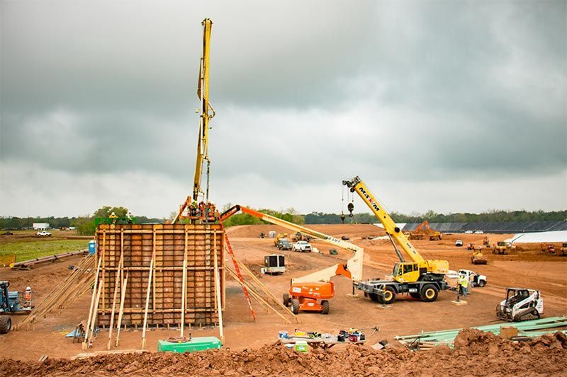 Construction site with cranes and workers pouring concrete beside a large foundation under cloudy skies