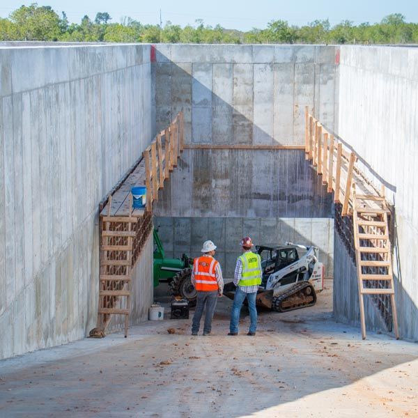 Workers stand in a concrete trench beside a compact excavator and wooden stairs on both sides.